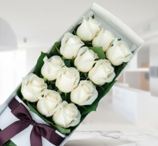 Caja de 12 rosas blancas frescas y abiertas con un moño elegante, sobre una superficie de mármol en un entorno de floristería