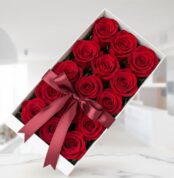 Caja blanca rectangular con doce rosas rojas frescas y abiertas, adornada con un elegante moño de satén color vino, sobre una