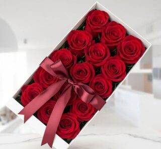 Caja blanca rectangular con doce rosas rojas frescas y abiertas, adornada con un elegante moño de satén color vino, sobre una