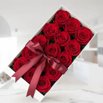 Caja blanca rectangular con doce rosas rojas frescas y abiertas, adornada con un elegante moño de satén color vino, sobre una