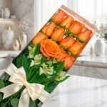 Caja de 12 rosas salmón frescas y abiertas con follaje verde y un moño elegante, sobre una superficie de mármol en un entorno