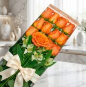 Caja de 12 rosas salmón frescas y abiertas con follaje verde y un moño elegante, sobre una superficie de mármol en un entorno