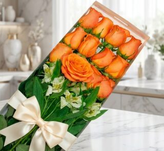 Caja de 12 rosas salmón frescas y abiertas con follaje verde y un moño elegante, sobre una superficie de mármol en un entorno