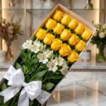 Caja de 18 rosas amarillas frescas y lirios blancos con lazo de satén blanco, sobre superficie de mármol, fondo de floristerí