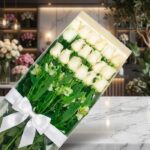 Caja transparente con 18 rosas blancas frescas y alstroemerias, atada con un moño blanco, sobre una superficie de mármol en u