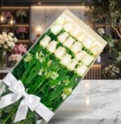 Caja transparente con 18 rosas blancas frescas y alstroemerias, atada con un moño blanco, sobre una superficie de mármol en u