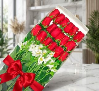 Caja transparente con 18 rosas rojas frescas y alstroemerias blancas, atada con un elegante moño rojo, sobre una superficie d