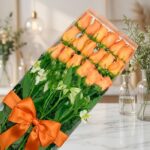 Caja transparente con 18 rosas salmón frescas y abiertas, acompañadas de follaje verde y flores blancas, atada con un moño na