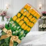 Caja transparente con 24 rosas amarillas vibrantes y alstroemerias blancas, atada con un moño dorado, sobre mármol blanco en