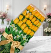 Caja transparente con 24 rosas amarillas vibrantes y alstroemerias blancas, atada con un moño dorado, sobre mármol blanco en