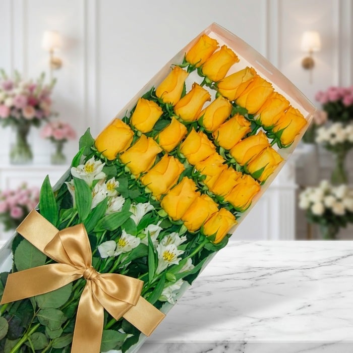 Caja transparente con 24 rosas amarillas vibrantes y alstroemerias blancas, atada con un moño dorado, sobre mármol blanco en
