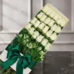 Caja de 24 rosas blancas frescas y abiertas, con follaje verde y un moño verde esmeralda, sobre una superficie de mármol oscu