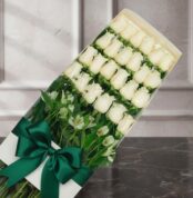 Caja de 24 rosas blancas frescas y abiertas, con follaje verde y un moño verde esmeralda, sobre una superficie de mármol oscu