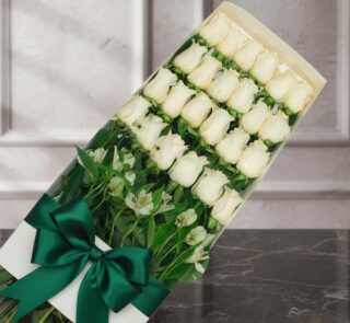 Caja de 24 rosas blancas frescas y abiertas, con follaje verde y un moño verde esmeralda, sobre una superficie de mármol oscu