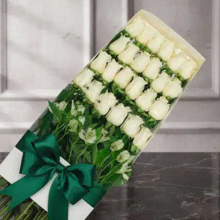 Caja de 24 rosas blancas frescas y abiertas, con follaje verde y un moño verde esmeralda, sobre una superficie de mármol oscu