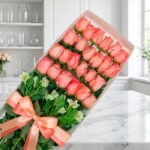 Caja de 24 rosas rosadas frescas y abiertas con follaje verde, atada con un moño de satén color salmón, sobre una superficie