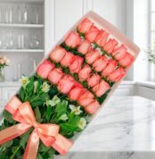 Caja de 24 rosas rosadas frescas y abiertas con follaje verde, atada con un moño de satén color salmón, sobre una superficie