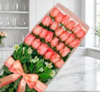 Caja de 24 rosas rosadas frescas y abiertas con follaje verde, atada con un moño de satén color salmón, sobre una superficie