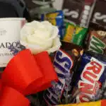 Primer plano extremo de una rosa blanca con gotas de rocío, lavanda, lazo rojo, chocolates Snickers y Run, y botella de vino.