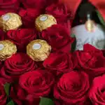 Primer plano extremo de la Caja Floral ENRICO con rosas rojas frescas, chocolates Ferrero Rocher y una botella de vino, desta