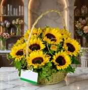 Canasta floral de mimbre con 12 girasoles grandes y vibrantes, follaje verde exuberante, sobre una superficie de mármol pulid