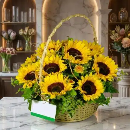 Canasta floral de mimbre con 12 girasoles grandes y vibrantes, follaje verde exuberante, sobre una superficie de mármol pulid