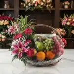 Canasta Frutal CARMINE con lirios rosados, gerberas fucsia, follaje verde y frutas frescas como uvas, manzanas, naranjas y ki