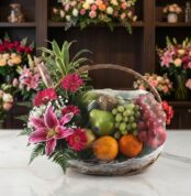 Canasta Frutal CARMINE con lirios rosados, gerberas fucsia, follaje verde y frutas frescas como uvas, manzanas, naranjas y ki