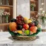 Canasta de frutas Corvus con uvas rojas, manzanas, kiwis y duraznos, sobre una superficie de mármol en un entorno de floriste