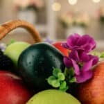 Primer plano extremo de una canasta con frutas frescas y flores moradas, destacando texturas y gotas de rocío.