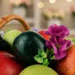 Primer plano extremo de una canasta con frutas frescas y flores moradas, destacando texturas y gotas de rocío.