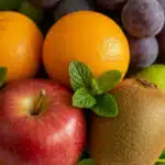 Primer plano macro de la Canasta de Frutas Draco, mostrando manzanas rojas, naranjas, kiwis, uvas y hojas de menta con gotas