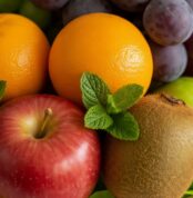 Primer plano macro de la Canasta de Frutas Draco, mostrando manzanas rojas, naranjas, kiwis, uvas y hojas de menta con gotas