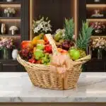 Canasta de mimbre con frutas gourmet variadas: piñas, uvas, manzanas, peras, mangos y plátanos, sobre mármol blanco en floris