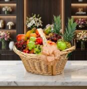 Canasta de mimbre con frutas gourmet variadas: piñas, uvas, manzanas, peras, mangos y plátanos, sobre mármol blanco en floris