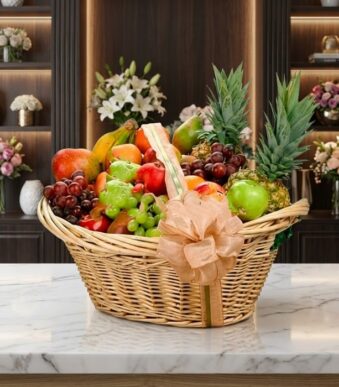 Canasta de mimbre con frutas gourmet variadas: piñas, uvas, manzanas, peras, mangos y plátanos, sobre mármol blanco en floris