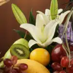 Primer plano extremo de una canasta de frutas premium con lirios blancos, kiwi, uvas, manzana, pera, limón y mango, con gotas