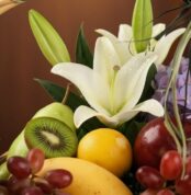 Primer plano extremo de una canasta de frutas premium con lirios blancos, kiwi, uvas, manzana, pera, limón y mango, con gotas