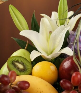 Primer plano extremo de una canasta de frutas premium con lirios blancos, kiwi, uvas, manzana, pera, limón y mango, con gotas