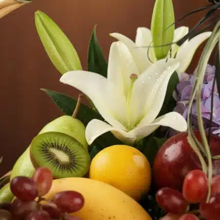 Primer plano extremo de una canasta de frutas premium con lirios blancos, kiwi, uvas, manzana, pera, limón y mango, con gotas
