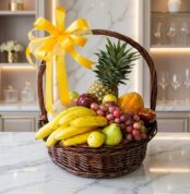 Canasta de mimbre con frutas frescas variadas como piña, plátanos, uvas, manzanas y peras, adornada con un lazo amarillo bril