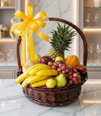 Canasta de mimbre con frutas frescas variadas como piña, plátanos, uvas, manzanas y peras, adornada con un lazo amarillo bril
