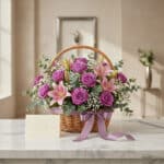 Cesta de flores AZURE con rosas moradas, lirios rosas y follaje verde, sobre una mesa de mármol con una tarjeta de regalo y c