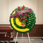 Corona fúnebre Adela con rosas rojas y flores amarillas, cinta morada 'En Profunda Condolencia', junto a un ataúd en sala de