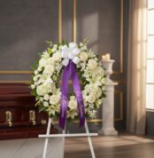 Corona fúnebre Almudena con flores blancas, cinta morada 'En Recuerdo Amoroso', junto a un ataúd en sala de velación premium.