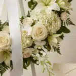 Fotografía macro premium de la Corona Fúnebre Almudena, con rosas, lirios y orquídeas blancas, follaje verde, rocío sutil y c