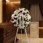 Corona de flores fúnebres Ararat con rosas rojas, lirios blancos y margaritas, junto a un ataúd de madera en un ambiente eleg