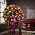 Corona fúnebre Atalia con rosas blancas, naranjas y rosadas, con cinta morada 'En Memoria Eterna' junto a un ataúd en sala de