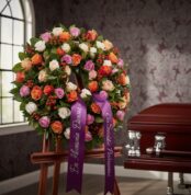 Corona fúnebre Atalia con rosas blancas, naranjas y rosadas, con cinta morada 'En Memoria Eterna' junto a un ataúd en sala de