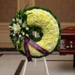Corona fúnebre Bethesda con flores blancas y amarillas, cinta morada con mensaje 'En Profunda Simpatía', junto a un ataúd en